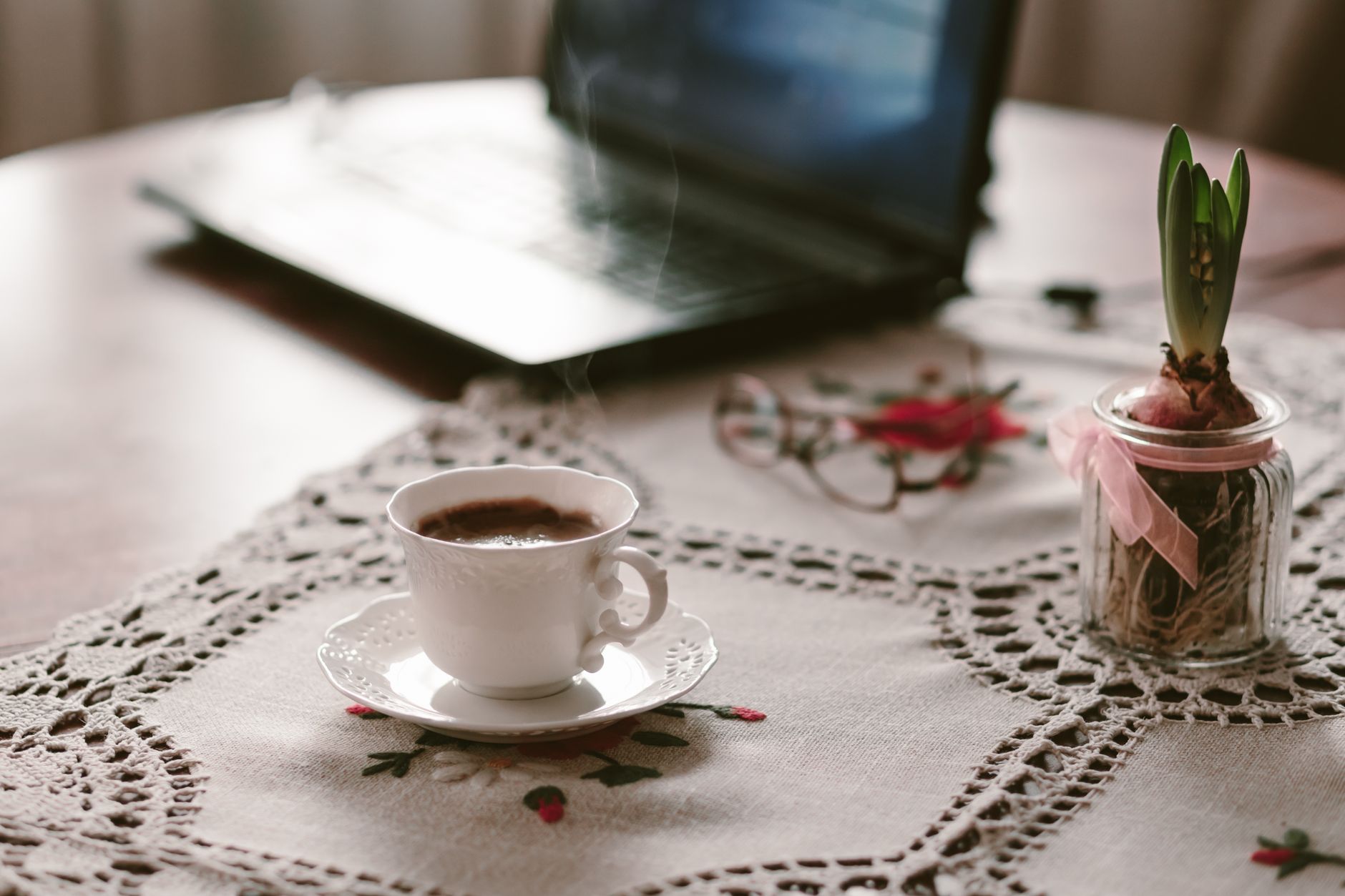 Un espacio de trabajo cálido y acogedor con una taza de café humeante junto a un portátil sobre un mantel de encaje.