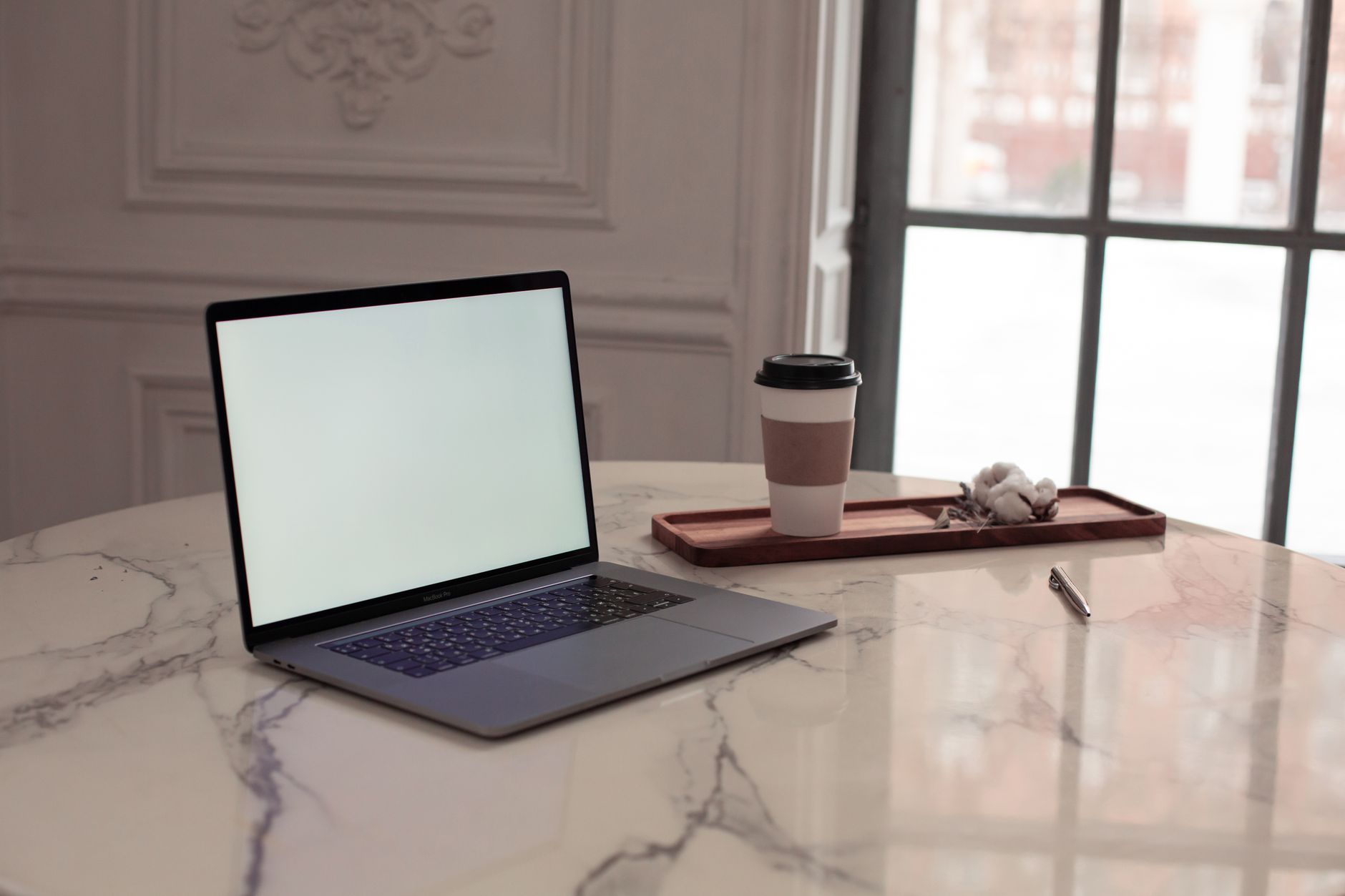 Una mesa minimalista con un portátil, una taza de café y un bolígrafo junto a una ventana.